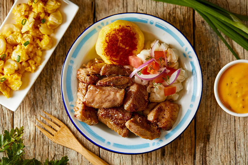 Auf einem rustikalen Holztisch steht ein Teller mit gebratenen Fleischstücken, einem goldbraunen Maispuffer, Zwiebel-Tomaten-Salsa und Mote. Ecuador Essen Fritada