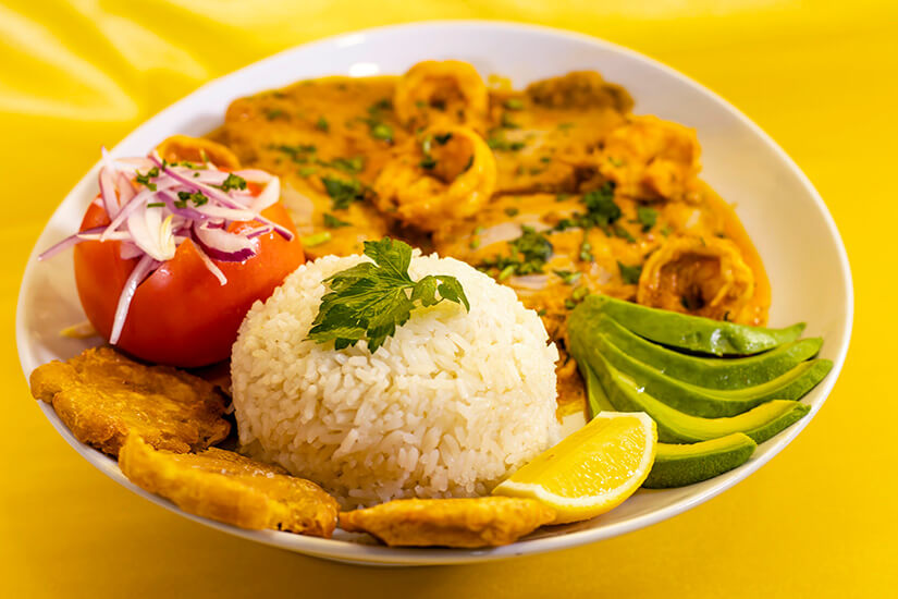 Auf einem leuchtend gelben Tischtuch steht ein Teller mit Meeresfrüchten in cremiger Kokossoße, Tintenfischringen und Garnelen, dazu eine Portion Reis, Avocado, frittierte Kochbananen, eine gefüllte Tomate mit roten Zwiebeln und eine Zitronenspalte. Encocado de Pescado
