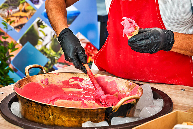 Ein Verkäufer mit roter Schürze und schwarzen Handschuhen rührt in einem großen Kupferkessel auf Eis pinkfarbenes Fruchteis und füllt es gerade in eine Waffeltüte. Ecuador Essen Eis
