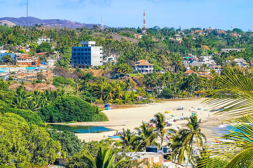 Ein heller Sandstrand vor der Stadt Puerto Escondido ist zu sehen. Palmen und viel Grün umgeben den Abschnitt. Im Hintergrund stehen einige Gebäude. Puerto Escondido Mexiko Willkommen