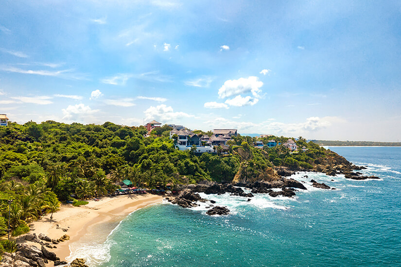 Eine Bucht gesäumt von grünem Dschungel ist zu sehen. Heller Sandstrand geht in türkisblaues Meer über. Puerto Escondido Mexiko Strand