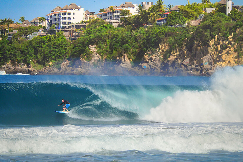 Ein Surfer reitet eine große Welle entlang. Im Hintergrund ist eine grün bewachsene Küste zu sehen, auf der weiße Gebäude thronen. Puerto Escondido Mexiko Surfen