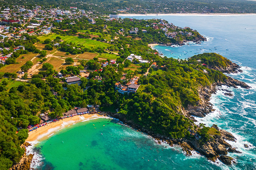 Puerto Escondido von oben betrachtet. Grüne Vegetation trifft auf hellen Sandstrand. Das Meer davor leuchtet wie ein Smaragd. Puerto Escondido Mexiko Wetter