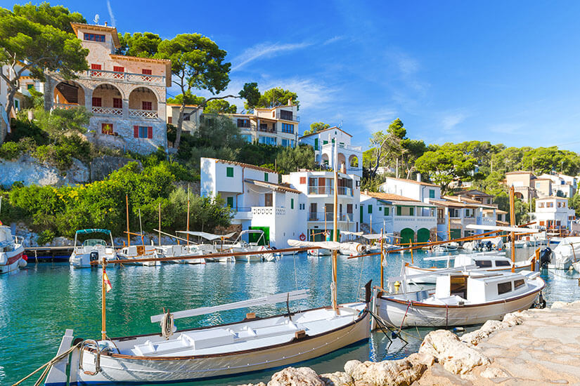 Ein idyllischer Fischerhafen in einer Bucht mit smaragdgrünem Wasser, an dessen Ufer traditionelle weiße Fischerboote vertäut sind, umgeben von mediterranen Häusern an einem bewaldeten Hang unter blauem Himmel. Menorca oder Mallorca Cala Figuera