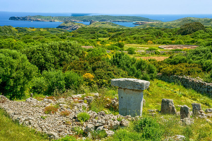 Eine historische Steinstruktur mit einer großen, flachen T-förmigen Taula-Steinformatiom im Vordergrund, umgeben von grünen, hügeligen Feldern, Trockenmauern und mediterraner Vegetation mit Blick auf das blaue Meer im Hintergrund. Menorca oder Mallorca Talayot
