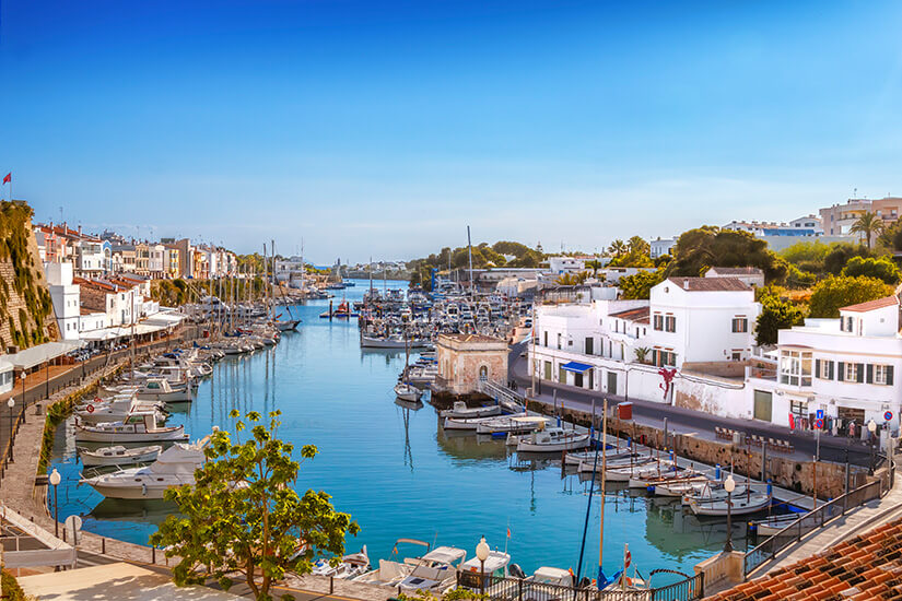 Ein langgestreckter Naturhafen in Ciutadella mit ruhigem, blauem Wasser, an dessen Ufern zahlreiche Boote und Jachten anlegen, flankiert von weißen Gebäuden, einer Uferpromenade und Bäumen unter einem strahlend blauen Himmel. Menorca oder mallorca Ciutadella