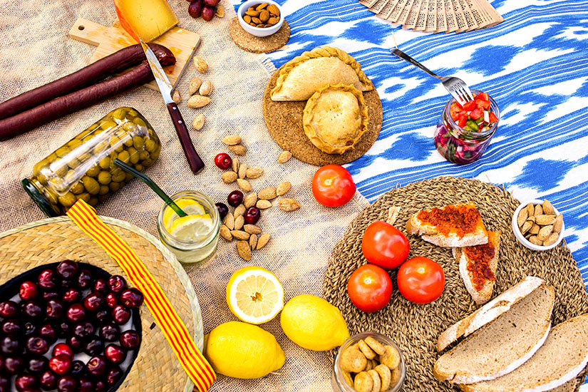 Ein reich gedecktes Picknick aus der Vogelperspektive auf einer Decke mit typischen Spezialitäten wie Sobrassada-Broten, Oliven im Glas, Tomaten, Käse, Gebäck, Mandeln, Kirschen und Zitronen neben einem Strohhut. Menorca oder Mallorca Picknick Majorcan