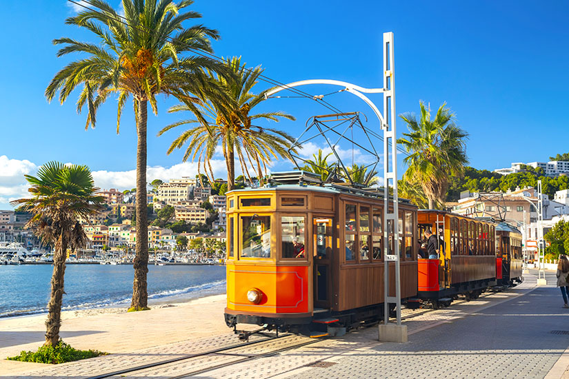 Eine historische, orange-braune Holzstraßenbahn fährt auf Schienen direkt an der Strandpromenade von Port de Sóller entlang, gesäumt von hohen Palmen mit Blick auf die Bucht und die hügelige Küstenstadt unter blauem Himmel. Menorca oder Mallorca Tram Port de Soller