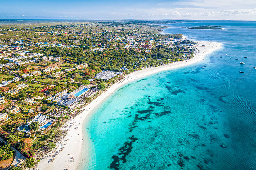 Kendwa zählt zu den Traumstränden im Nordwesten Sansibars und gilt als einer der wenigen ohne starke Gezeiten. Eine breite, geschwungene Bucht mit puderfeinem Sand wird von eleganten Resorts und tropischem Grün gesäumt. Im klaren Wasser zeichnen sich Riffstrukturen ab, am Horizont ankern Segelboote. Sansibar Strände Kendwa Beach
