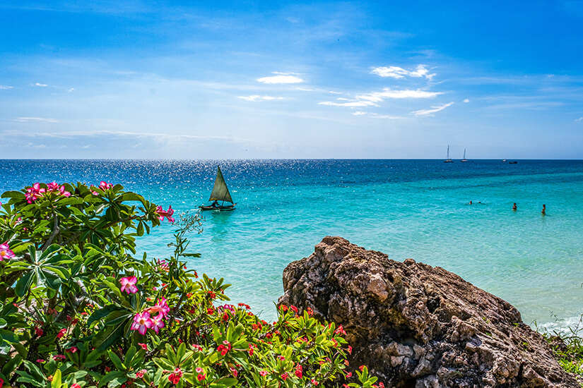 Nungwi an der Nordspitze Sansibars gilt als bester Strand auf Sansibar zum Schwimmen. Tropische Blüten und Felsen rahmen den Blick auf das kristallklare Meer. Traditionelle Dhau-Segelboote und moderne Yachten kreuzen das türkise Wasser. Sansibar Strände Nungwi Beach