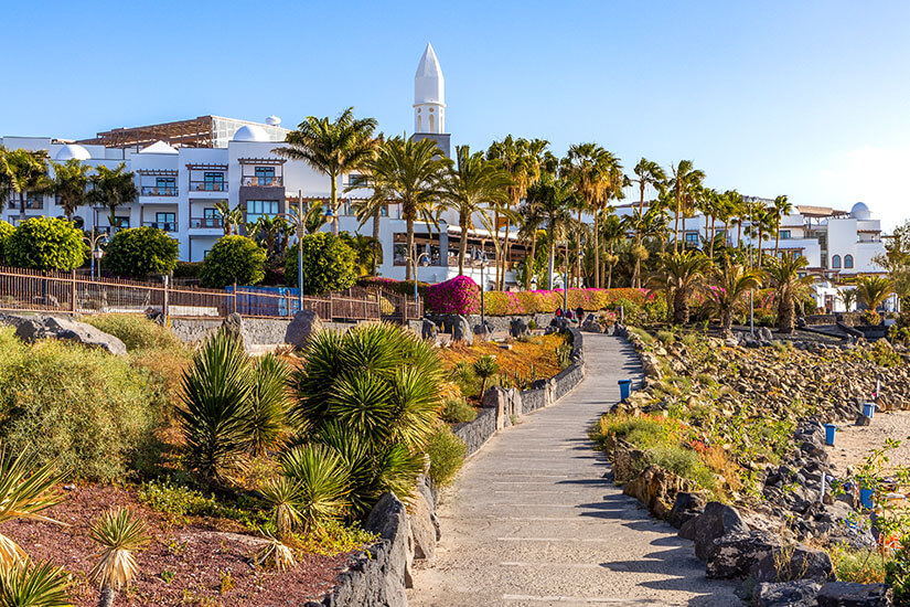 Eine gepflasterte Strandpromenade schlängelt sich entlang einer steinigen Küste, gesäumt von grünen Sukkulenten, Palmen und blühenden lila Büschen vor einer großen, weißen Hotelanlage mit einem markanten weißen Turm unter strahlend blauem Himmel. Playa Blanca Spanien Promenade