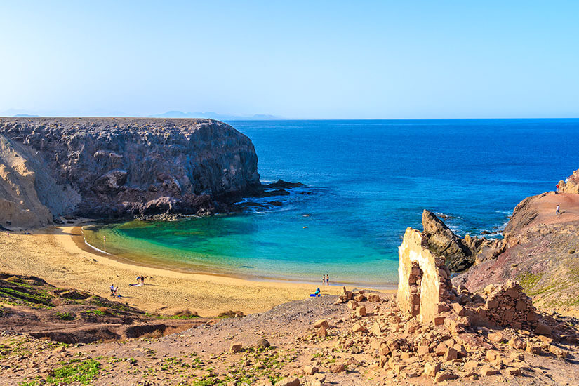 Ein Blick von oben auf eine traumhafte, halbkreisförmige Badebucht mit feinem, goldenem Sandstrand und glasklarem, smaragdgrünem bis tiefblauem Wasser, geschützt von hohen, dunklen Klippen, mit einer alten Steinruine im felsigen Vordergrund. Playa Blanca Spanien Playa de Papagayo