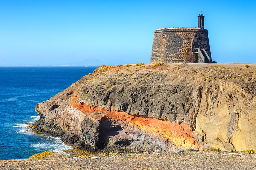 Ein runder, historischer Wehrturm aus dunklem Stein steht auf einer steilen Klippe mit markanten, rötlich und gelb geschichteten Gesteinsformationen, umspült vom tiefblauen Meer unter einem wolkenlosen Himmel. Playa Blanca Spanien Castillo de las Coloradas