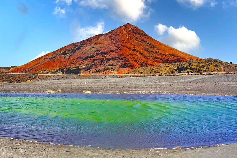 Landschaftsaufnahme im Timanfaya-Nationalpark auf Lanzarote, die eine auffällige, smaragdgrüne und dunkelviolette Lagune im Vordergrund zeigt. Die Lagune wird von dunklem vulkanischem Ascheufer begrenzt. Playa Blanca Spanien Timanfaya