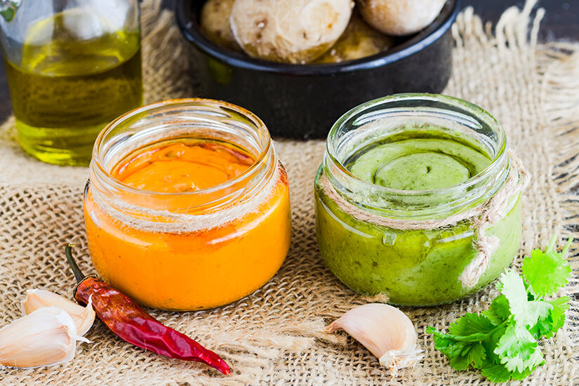 Zwei kleine Gläser, gefüllt mit roter und grüner Mojo-Soße, stehen nebeneinander auf einem groben Jutetuch, dekoriert mit Knoblauchzehen, einer getrockneten Chilischote und frischem Koriander, während im Hintergrund eine Schale mit runzeligen Kanarischen Kartoffeln (Papas Arrugadas) und eine Flasche Olivenöl zu sehen sind. Playa Blanca Spanien Papas Arrugadas