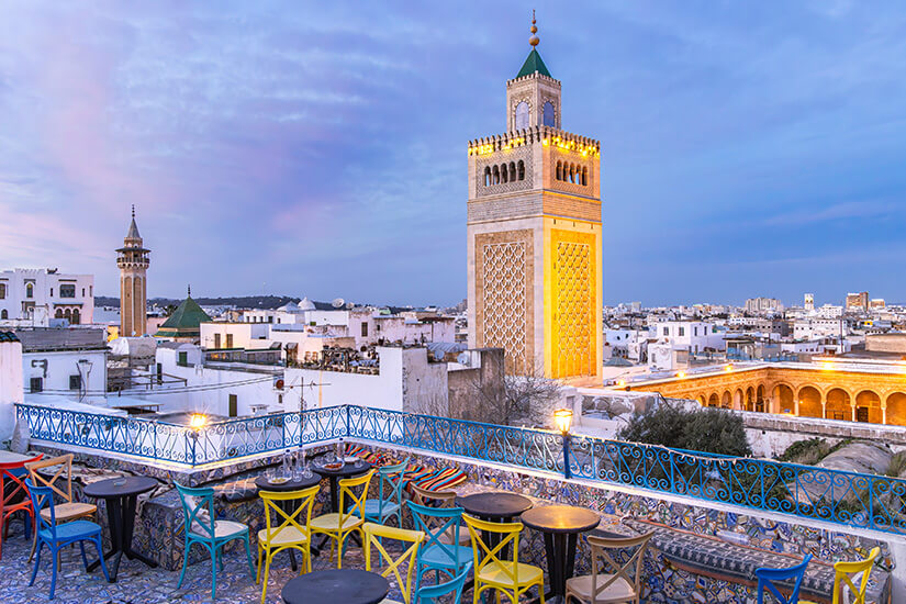 Von einer Dachterrasse mit bunten Stühlen und gefliesten Bänken blickt man in der Abenddämmerung über die weißen Häuser der Medina von Tunis auf das beleuchtete Minarett der Zitouna-Moschee. Tunesisches Essen Medina