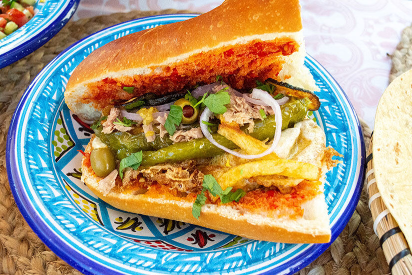 Auf einem blau gemusterten Keramikteller liegt ein üppig gefülltes Baguette-Sandwich mit Harissa, Thunfisch, gegrillten grünen Paprika, Oliven, roten Zwiebelringen, Pommes frites und frischen Kräutern. Tunesisches Essen Kafteji