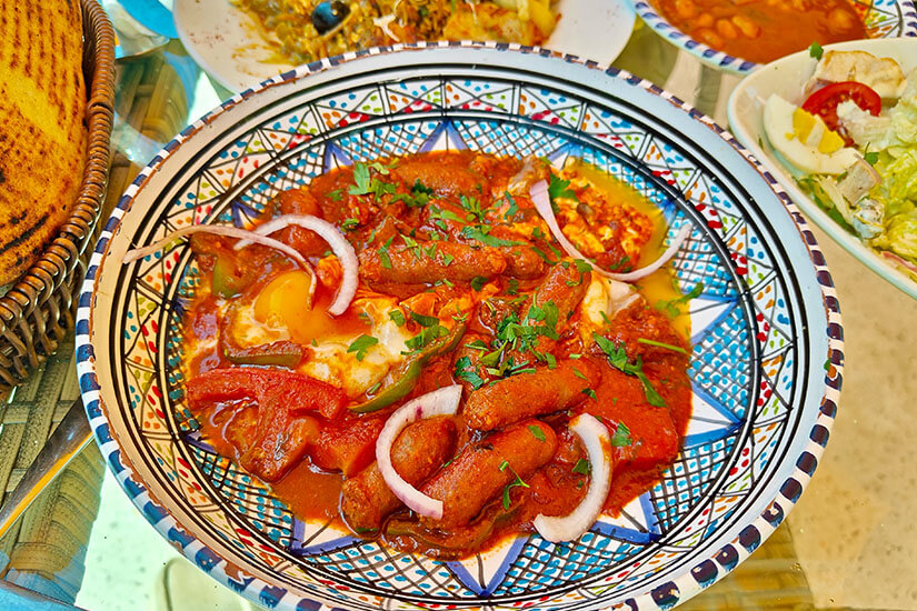 In einer farbenfroh gemusterten Keramikschale dampft ein würziges Gericht aus Merguez-Würstchen, pochierten Eiern, Paprika und roten Zwiebelringen in einer kräftigen Tomaten-Harissa-Soße. Tunesisches Essen Ojja