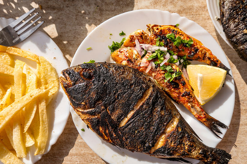 Auf einem weißen Teller liegen eine ganze gegrillte Dorade und ein aufgeklappter Fisch mit Harissa-Marinade, garniert mit roten Zwiebeln, Petersilie und einer Zitronenspalte, dazu eine Portion Pommes frites. Gegrillter Fisch Tunesien