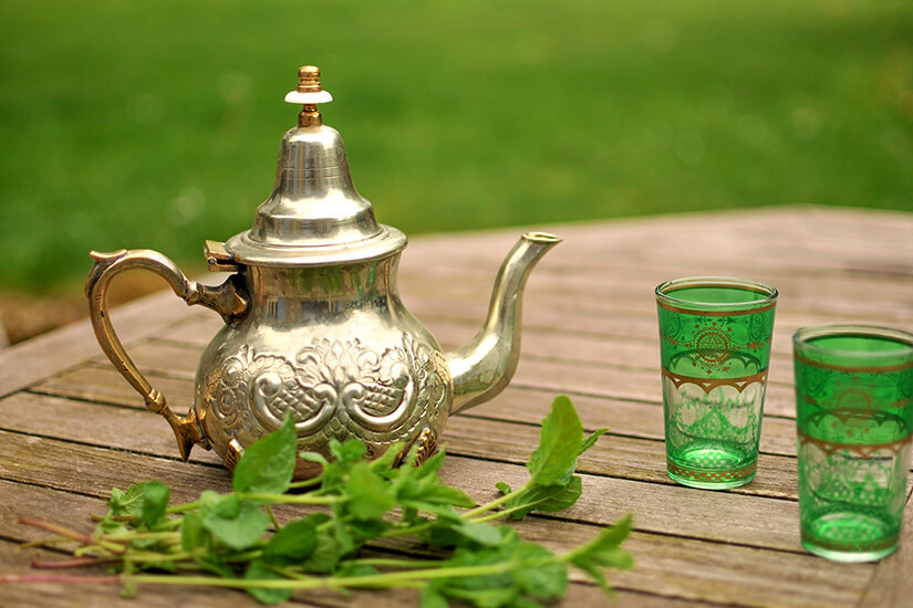 Auf einem Holztisch im Freien stehen eine kunstvoll verzierte silberne Teekanne, zwei grüne Teegläser mit goldenen Ornamenten und ein frischer Minzezweig bereit für die Zubereitung von nordafrikanischem Tee. Tunesisches Essen Tee