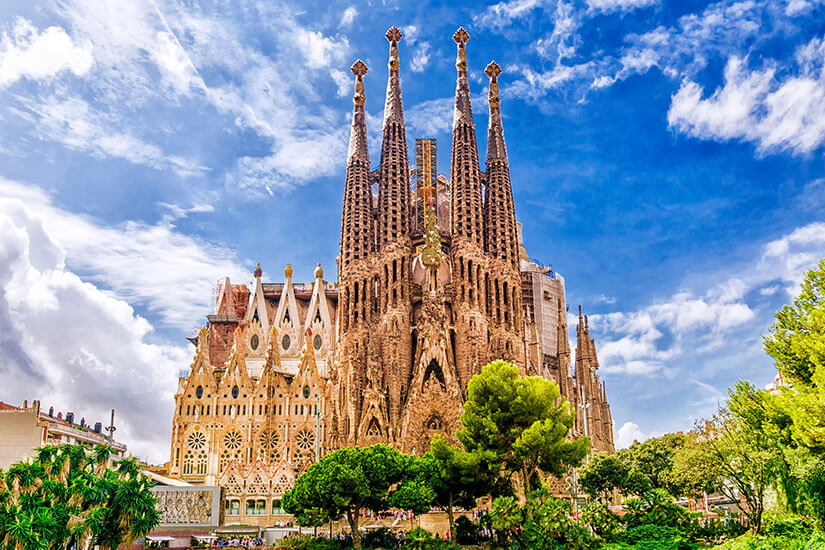 Vor einem leuchtend blauen Himmel mit Schäfchenwolken ragen die filigranen Türme der Sagrada Família in Barcelona empor, umgeben von grünen Bäumen. Wochenendtrip Barcelona