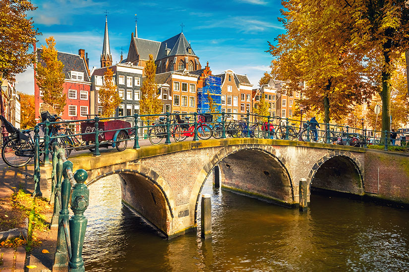 Eine charakteristische gemauerte Bogenbrücke mit zahlreichen abgestellten Fahrrädern überspannt einen Amsterdamer Grachtenkanal, gesäumt von typischen Giebelhäusern und herbstlich gefärbten Bäumen unter blauem Himmel. Wochenendtrip Amsterdam