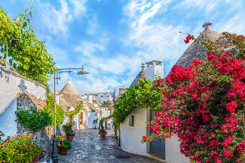 Malerische Gasse mit weißen Trulli-Häusern, üppigem Grün und leuchtenden Bougainvillea-Blüten. Das Bild vermittelt typisch apulisches Flair und eine warme, mediterrane Atmosphäre. Trulli, Gassen und Apulien-Feeling
