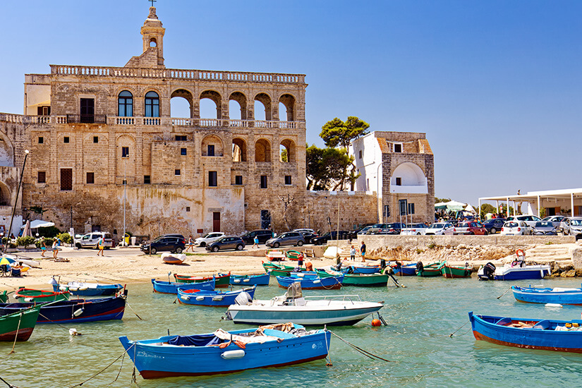 Bunte Fischerboote liegen vor historischen Gebäuden direkt am Wasser. Die Küstenstadt wirkt charmant, lebendig und typisch süditalienisch. Apulien oder Kalabrien Polignano