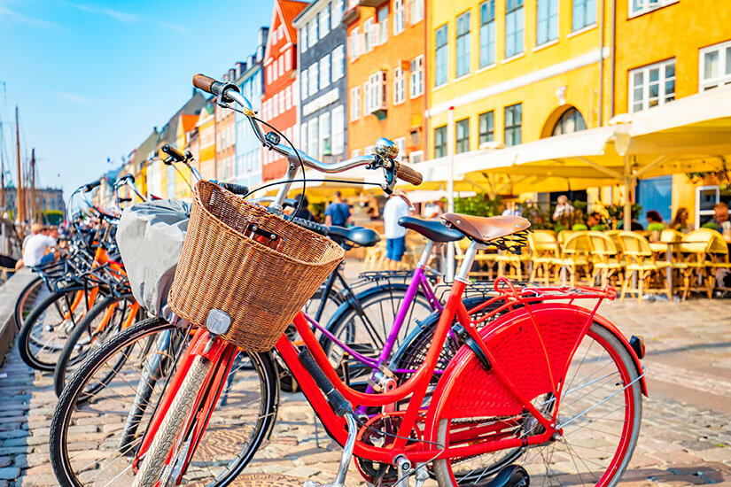 Im Vordergrund stehen abgestellte Fahrräder mit Korb am Kopfsteinpflaster, während sich im Hintergrund die berühmten bunten Giebelhäuser und Straßencafés am Hafen von Nyhavn in Kopenhagen aufreihen. Wochenendtrip Kopenhagen