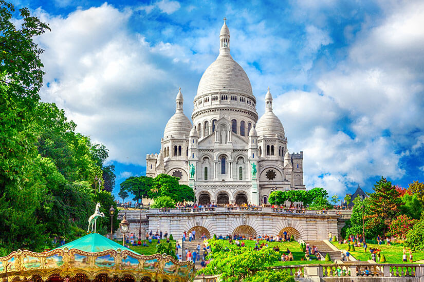 Vor einem dramatischen Wolkenhimmel thront die strahlend weiße Basilika Sacré-Cœur auf dem Hügel von Montmartre, umrahmt von grünen Bäumen, gepflegten Grünflächen mit Besuchern und einem historischen Karussell im Vordergrund. Wochenendtrip Paris