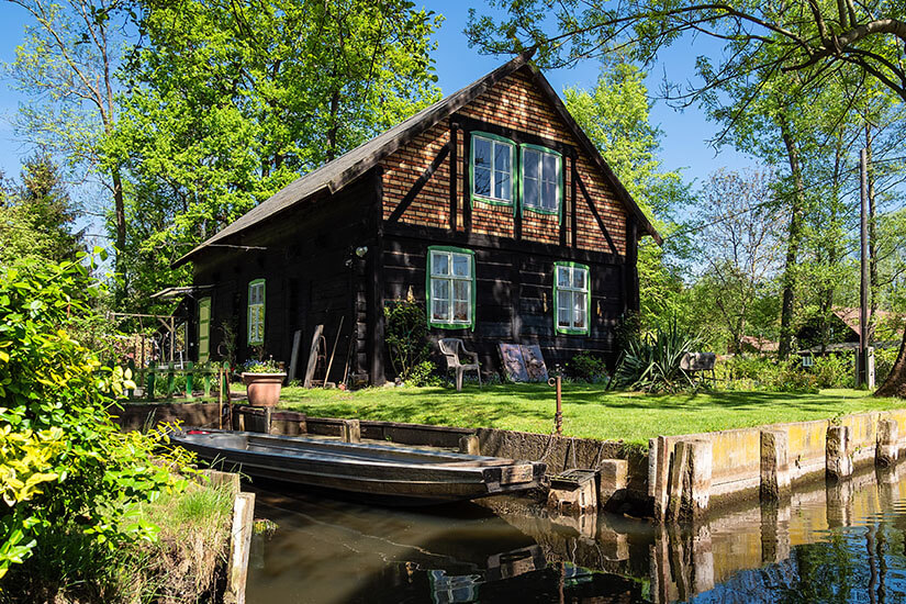Ein traditionelles dunkles Holzhaus mit grünen Fensterrahmen und Fachwerkgiebel steht idyllisch an einem kleinen Wasserkanal, an dessen Anlegestelle ein typischer flacher Holzkahn vertäut ist. Wochenendtrip Spreewald