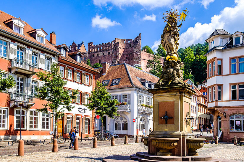 Ein gepflasterter Platz, der Kornmarkt in Heidelberg, mit dem barocken Madonna-Brunnen im Vordergrund, flankiert von eleganten historischen Stadthäusern. Hoch über den Dächern thront im Hintergrund die imposante Ruine des Heidelberger Schlosses am bewaldeten Hang unter einem blauen Himmel mit weißen Wolken. Schoenste Orte Sueddeutschland Heidelberg