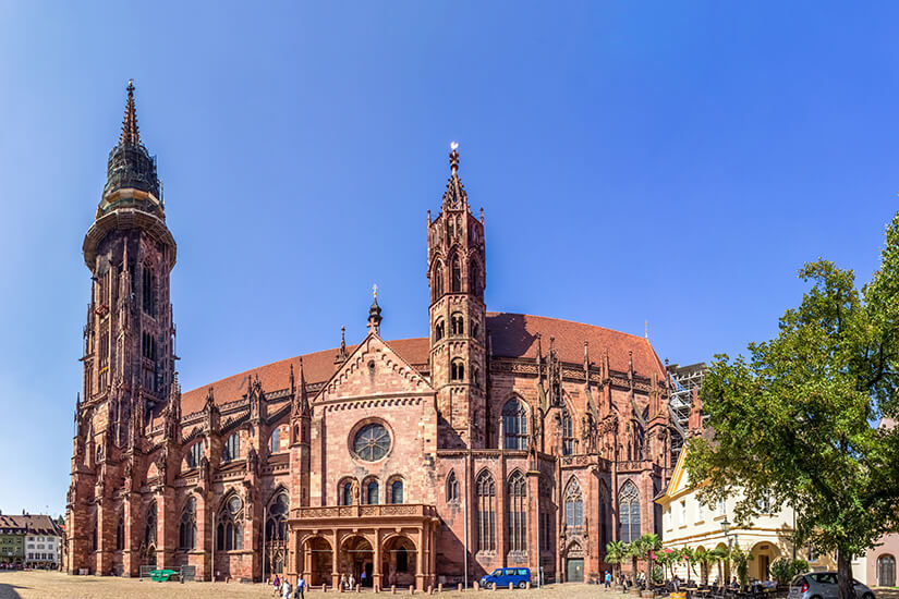 Die monumentale Außenansicht des Freiburger Münster aus rotem Sandstein zeigt die gotische Architektur des Kirchenschiffs im Detail unter einem wolkenlosen blauen Himmel. Schoenste Orte Sueddeutschland Freiburg