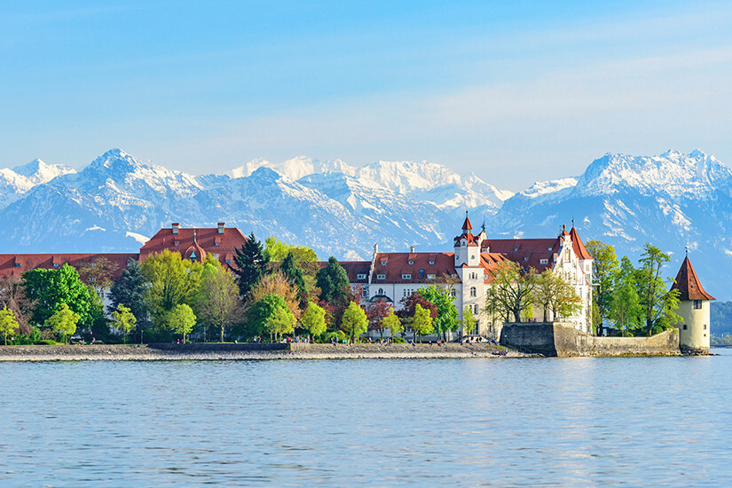 Eine weite Aufnahme des Bodensees zeigt die bayerische Stadt Lindau mit der historischen Insel und dem Schloss Montfort am Ufer, das von grünen Bäumen umgeben ist. Im Hintergrund erheben sich die schneebedeckten Gipfel der Schweizer Alpen. Schoenste Orte Sueddeutschland Bodensee