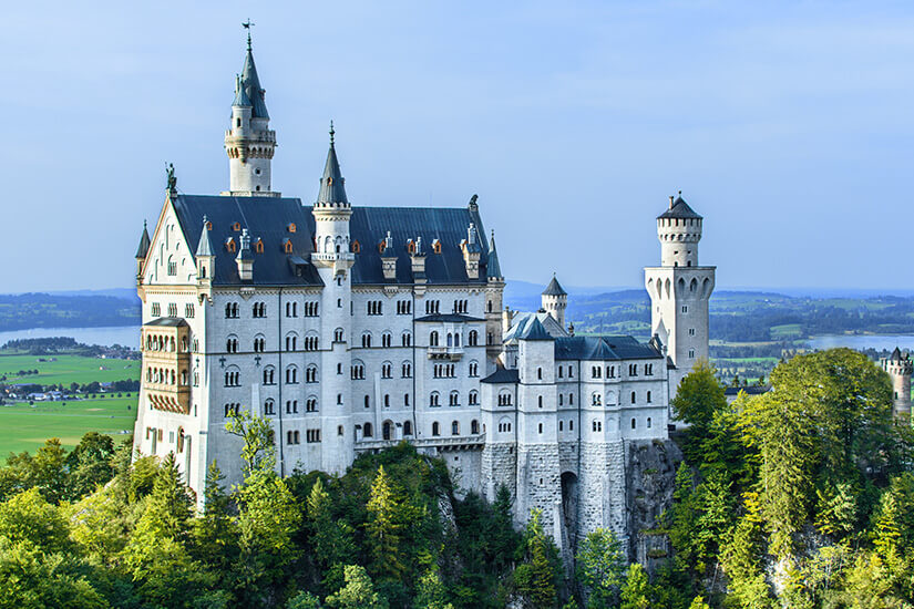 Das weltberühmte Schloss Neuschwanstein thront majestätisch auf einem bewaldeten Felsen und zeigt seine hellen Fassaden, spitzen Türme und dunklen Dächer im Detail. Schoenste Orte Sueddeutschland Neuschwanstein