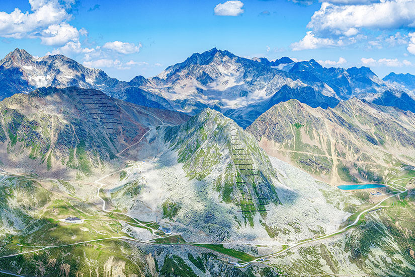 Eine beeindruckende Hochgebirgslandschaft mit schroffen, teils felsigen Gipfeln erstreckt sich bis zum Horizont. Serpentinenstraßen und Wege ziehen sich durch die Täler, während rechts ein kleiner türkisfarbener Bergsee zu sehen ist. Oetztal Region
