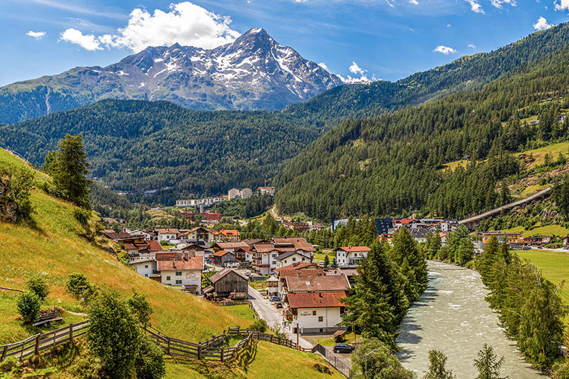 Ein Dorf mit traditionellen Häusern liegt in einem breiten Alpental, durch das ein Fluss verläuft. Dichte Nadelwälder bedecken die umliegenden Hänge, während im Hintergrund ein markanter, teils schneebedeckter Berggipfel aufragt. Oetztal Soelden