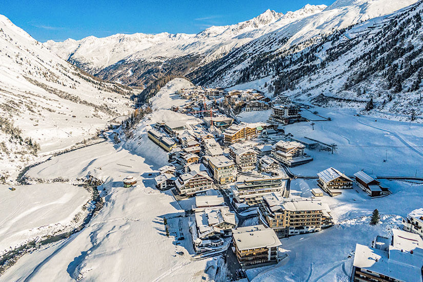Ein von Schnee bedeckter Wintersportort liegt eingebettet in einem engen Hochgebirgstal. Umgeben wird die Siedlung von verschneiten Hängen und hohen Berggipfeln, während sich ein Bach durch die winterliche Landschaft schlängelt. Oetztal Obergurgl