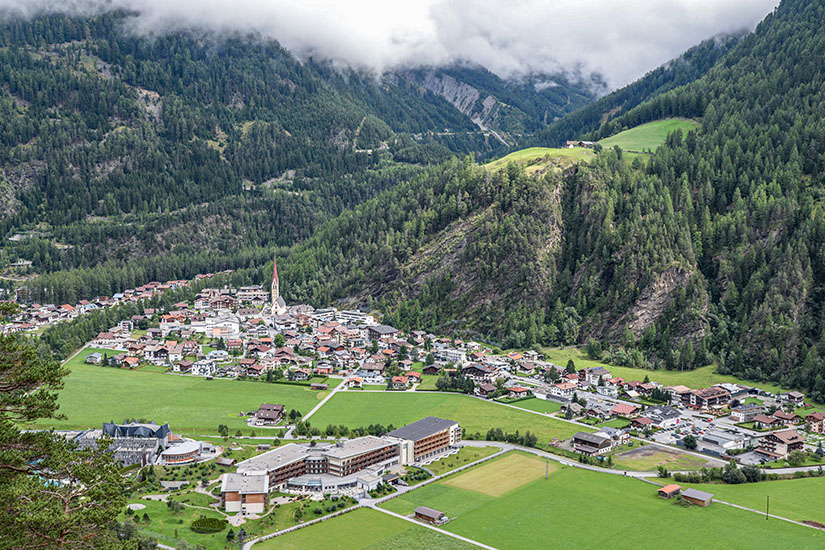 Ein Ort mit zahlreichen Häusern und einer markanten Kirche mit hohem Kirchturm liegt in einem grünen Alpental. Umgeben wird die Siedlung von bewaldeten Berghängen, über denen tiefe Wolken hängen. Oetztal Laengenfeld