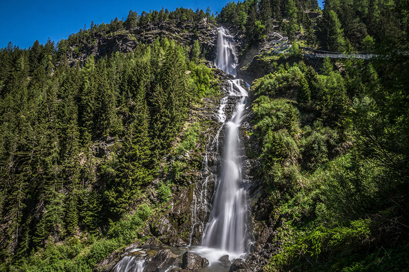 Ein hoher Wasserfall stürzt in mehreren Kaskaden über steile Felsen durch einen dichten Bergwald. Die umliegenden Hänge sind von Nadelbäumen und üppigem Grün bedeckt. Tirol Stuibenfall
