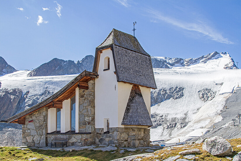 Eine kleine Kapelle aus Stein und Holz steht auf einer hochalpinen Landschaft vor einem ausgedehnten Gletscher. Dahinter erheben sich schroffe, teils schneebedeckte Berggipfel unter einem klaren Himmel. Tirol Rettenbachferner