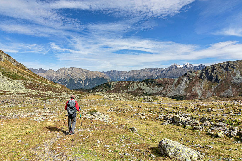 Eine Person mit Rucksack und Wanderstöcken bewegt sich durch eine weitläufige alpine Landschaft. Karge Wiesen, verstreute Felsen und schroffe Bergketten prägen die Umgebung bis zum Horizont. Tirol Wandern