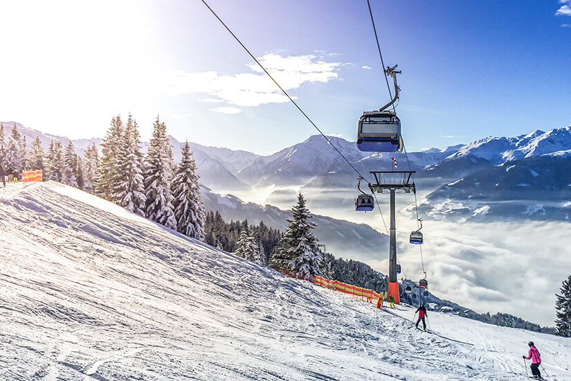 Eine verschneite Skipiste führt durch eine beeindruckende Berglandschaft mit Blick auf ein wolkenverhangenes Tal. Mehrere Gondeln schweben an einer Seilbahn über den Hang, während einige Skifahrer die Piste hinabfahren. Oetztal Skigebiet