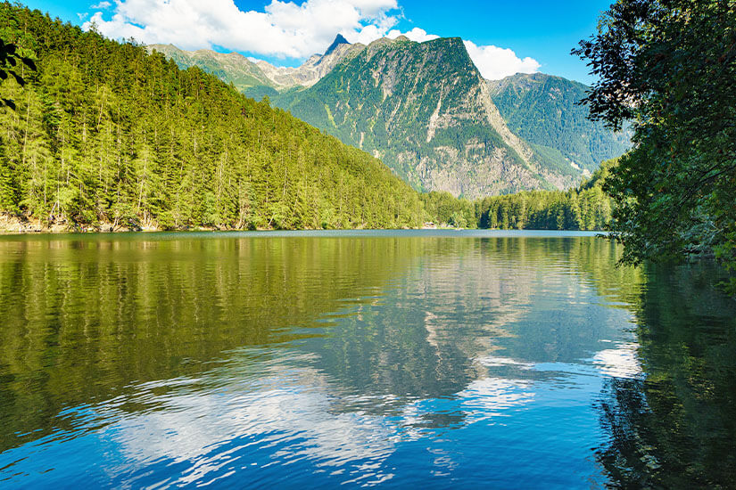 Ein ruhiger Bergsee liegt eingebettet zwischen dicht bewaldeten Hängen und hohen Bergen. Die Wasseroberfläche spiegelt die umliegenden Wälder und Gipfel wider. Tirol Piburger See