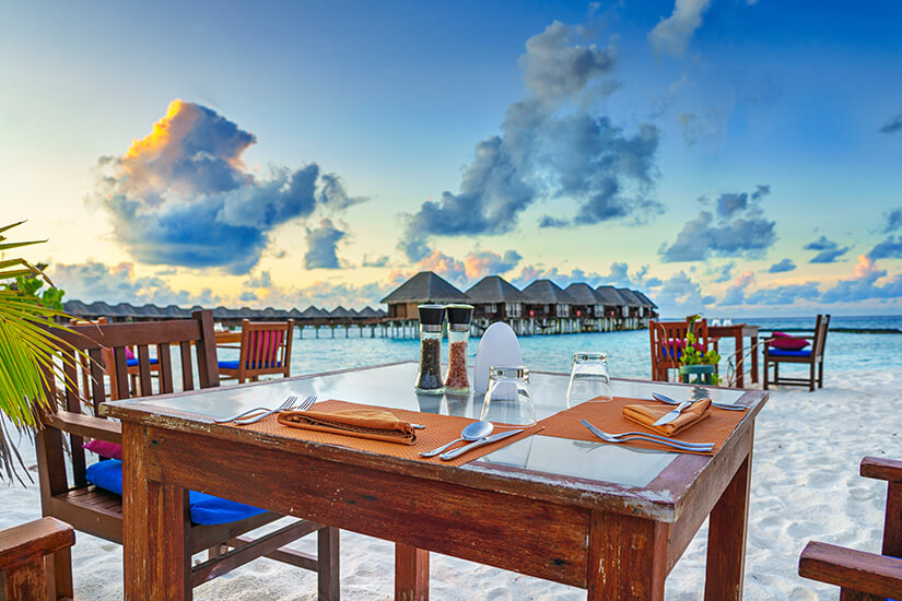 Ein liebevoll gedeckter Holztisch mit Besteck, Servietten und Gewürzmühlen steht im weißen Sand eines Strandrestaurants, im Hintergrund spiegeln sich die typischen Wasserbungalows der Malediven. Malediven Essen Restaurant