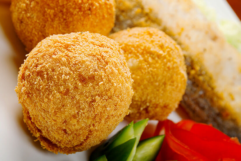 Auf einem Teller liegen goldbraun frittierte, panierte Bällchen mit knuspriger Hülle neben Gurken- und Tomatenscheiben sowie einem Stück gegrilltem Fisch. Malediven Essen Gulha