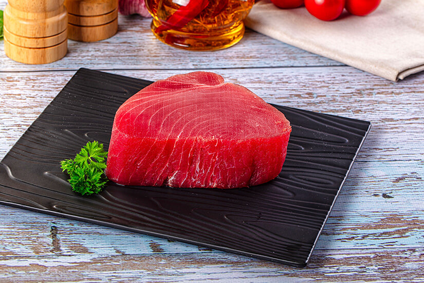 Auf einem schwarzen Schieferteller liegt ein dickes, leuchtend rotes Thunfischsteak, garniert mit frischer Petersilie, im Hintergrund sind Cherrytomaten, eine Karaffe Öl und Holzgewürzmühlen zu sehen. Malediven Essen Thunfisch