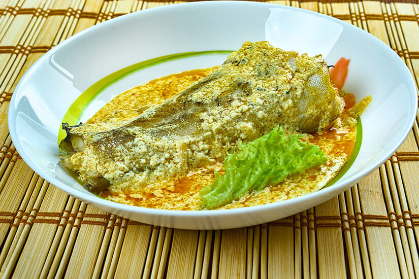 In einem weißen Teller mit grünem Rand liegt ein Fischfilet in einer cremig-orangefarbenen Kokos-Curry-Sauce, garniert mit krausem Salat und Tomatenstücken, präsentiert auf einer Bambusmatte. Mas Riha Essen