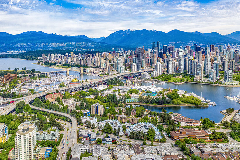 Eine Stadt mit Wolkenkratzer erstreckt sich vor einer Gebirgskette. Im Vordergrund spannt sich eine Brücke über einen Fluss und einige Grünflächen. Kanada Staedte Vancouver