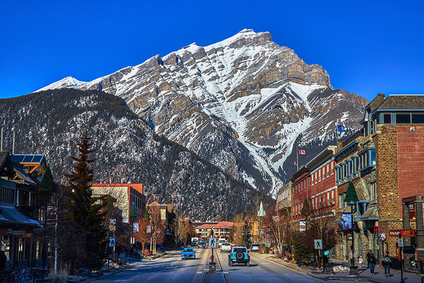 Eine gerade Hauptstraße führt durch einen Ort. Im Hintergrund ist ein schneebedeckter Gipfel zu sehen. Staedte Kanada Banff
